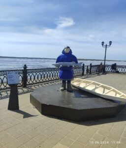 Фото памятника представителям народа Ханты. Мужчина в народной одежде народа Ханты держит в руках рыбу, позади него лежит лодка облас.