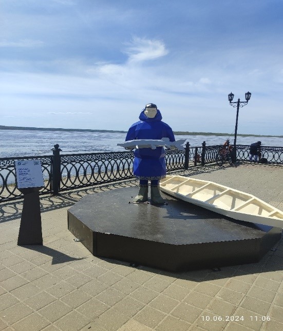 Фото памятника представителям народа Ханты. Мужчина в народной одежде народа Ханты держит в руках рыбу, позади него лежит лодка облас. 