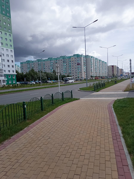 Фото: слева расположены многоэтажные дома, справа тротуар с   расположенным по двум сторонам от него газоном, посередине между домами и тротуаром с газоном - автомобильная дорога.  