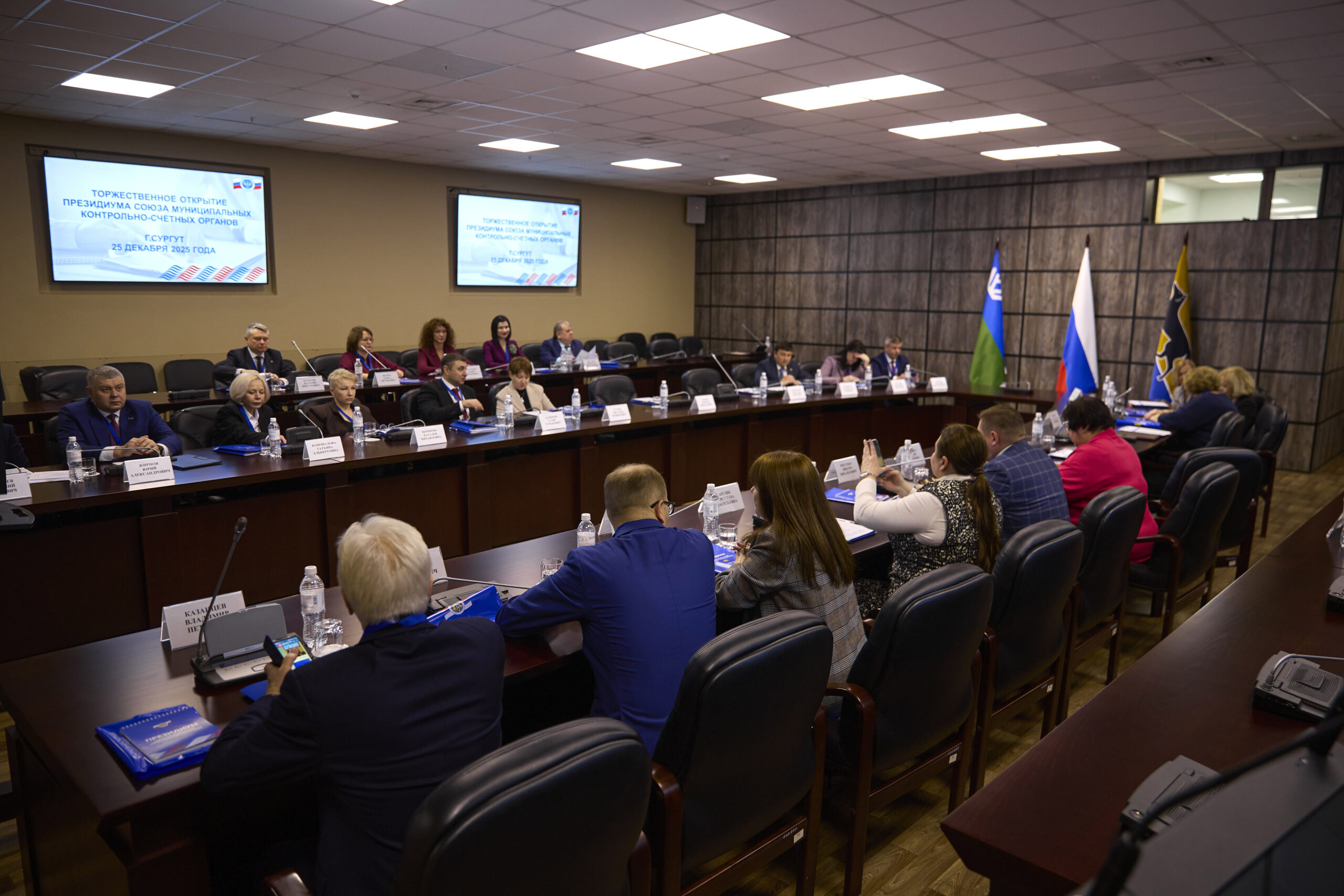 На фото — официальное заседание в конференц-зале. За длинным столом сидят около 20 человек в деловой одежде, они участвуют в обсуждении. Перед каждым участником — таблички с именами, микрофоны, бутылки с водой, документы.
На заднем плане — два больших экрана с текстом: «Торжественное открытие президиума Союза муниципальных контрольно-счётных органов. г. Сургут, 25 декабря 2025 года». Справа от зала видны три флага, среди них — флаг России.
Обстановка строгая: тёмные офисные кресла, деревянные столы, нейтральные стены. Освещение яркое, потолочные светильники равномерно распределены по потолку. Атмосфера официальная, деловая.