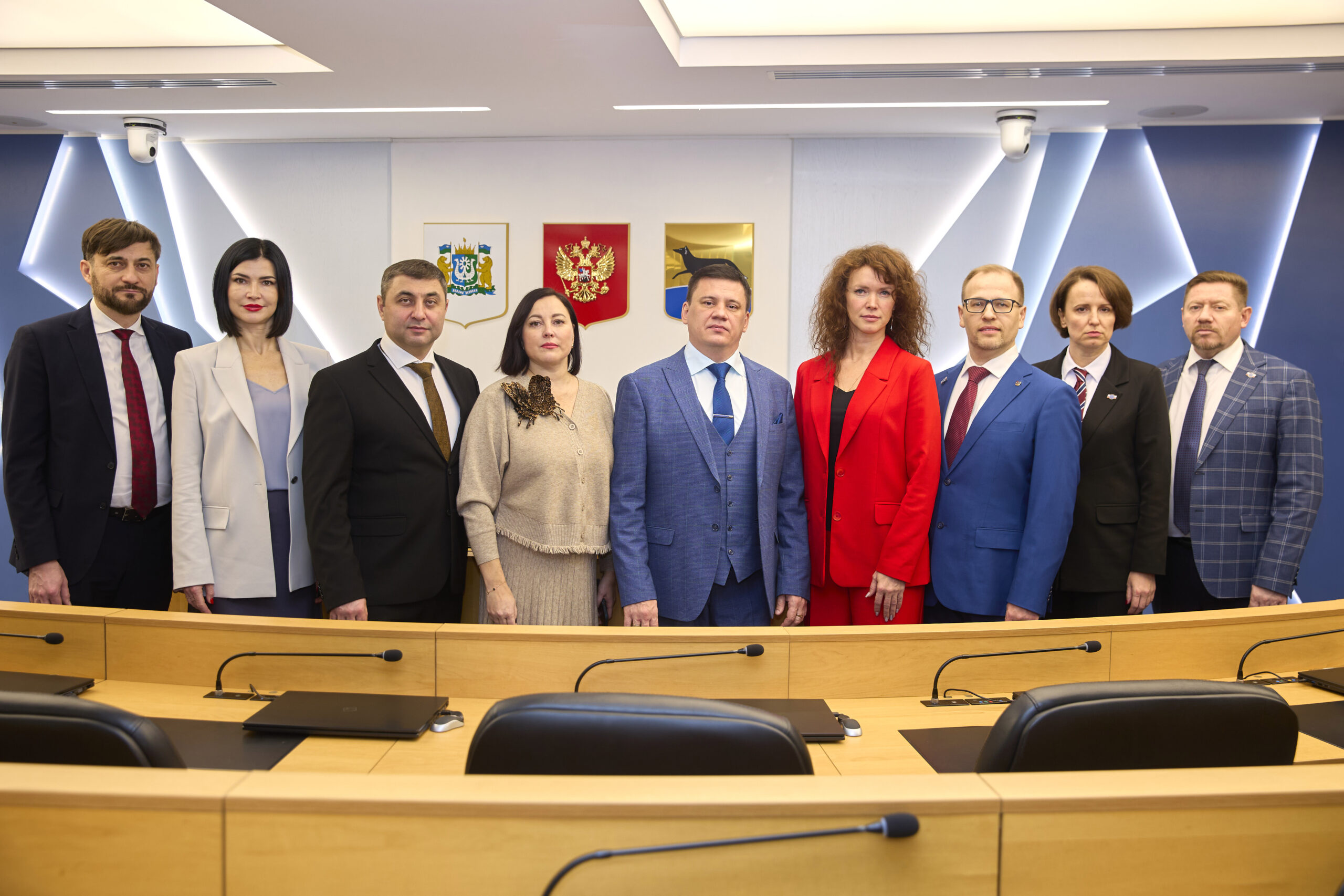 На фотографии изображена группа из девяти человек, стоящих в ряд в официальном помещении, похожем на зал заседаний. Все участники одеты в деловую одежду: мужчины — в костюмах с галстуками, женщины — в строгих платьях и жакетах. Среди одежды встречаются тёмные и светлые оттенки, выделяется ярко-красный жакет одной из женщин.

Люди стоят за длинным деревянным столом, на котором расположены микрофоны, ноутбуки и канцелярские принадлежности. Перед столом видны чёрные офисные кресла.

На заднем плане — стена с современным дизайном: геометрические световые элементы в синих и белых тонах. На стене размещены три герба: один из них — герб Российской Федерации (двуглавый орёл на красном фоне), два других — региональные или муниципальные символы.

В верхней части помещения видны потолочные светильники и камеры видеонаблюдения. Обстановка выглядит официально и строго, создаёт впечатление деловой или правительственной атмосферы.