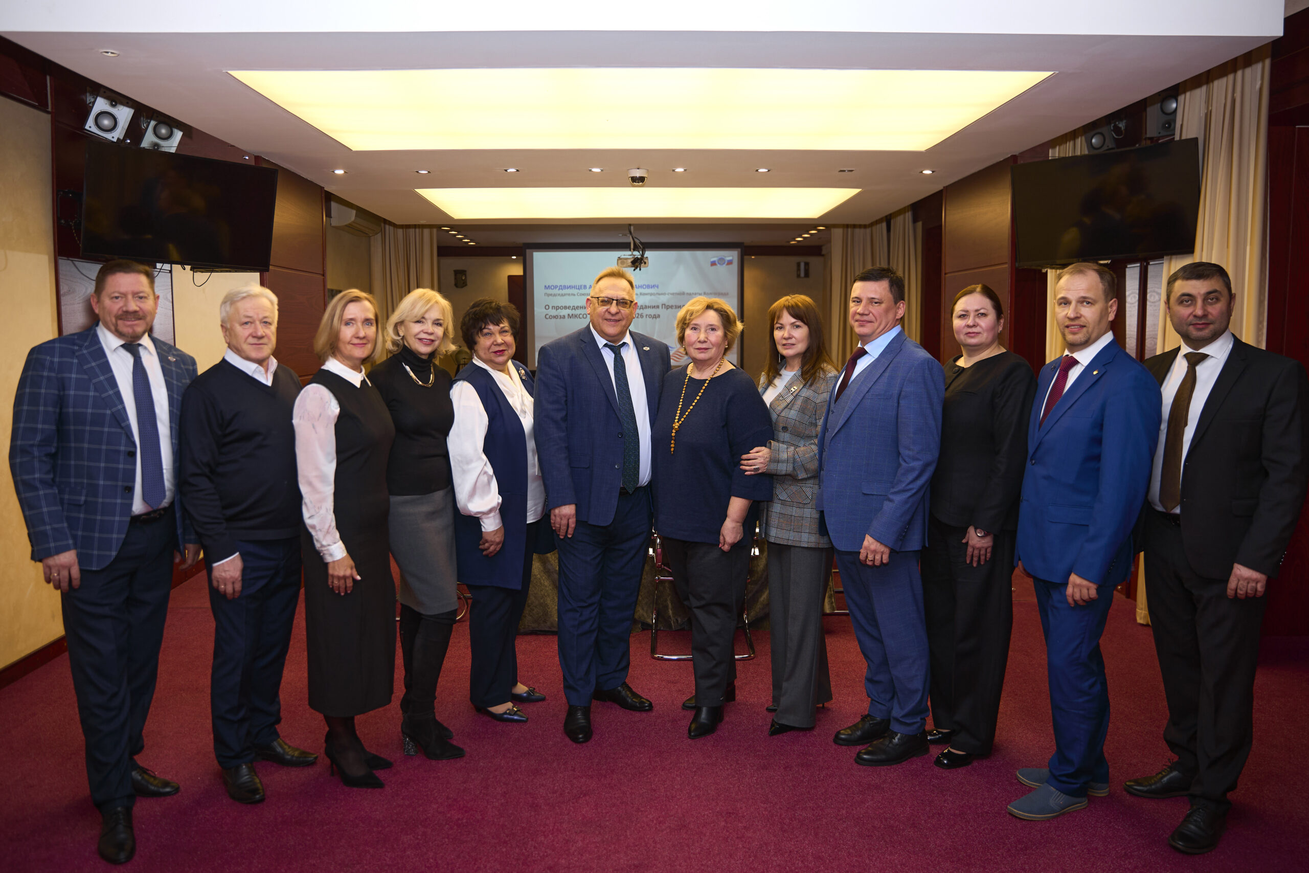На фото группа из 11 человек (мужчины и женщины) позирует для снимка в конференц-зале. Все одеты официально: мужчины — в костюмах с галстуками, женщины — в элегантных платьях и жакетах. Люди стоят в один ряд, улыбаются и смотрят в камеру.

Фон — интерьер зала с бордовыми стенами, шторами и ковровым покрытием бордового оттенка. На заднем плане виден экран с презентацией, на котором частично читается текст о заседании Союза МКСО. По бокам зала установлены большие телевизоры. Освещение яркое, потолочные светильники равномерно освещают помещение. Атмосфера официальная, деловая.