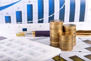 Фото-иллюстрация, на котором изображены: калькулятор, три стопки монет, ручка, расположенные на фоне листа, с изображённой на нем диаграммой. На первом плане: калькулятор, стопки монет и ручка. На заднем плане - лист с диаграммой.
