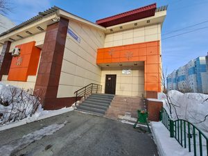 Фото входной зоны здания МКУ «Казна городского хозяйства». На изображении здание с бежевыми и оранжевыми панелями, чёрной дверью и лестницей. Вокруг снег, забор и кустарники.